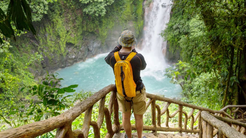 Person with a yellow backpack, perfect for what to pack for Costa Rica, taking a photo of a waterfall surrounded by lush greenery and turquoise water from a wooden viewing platform.